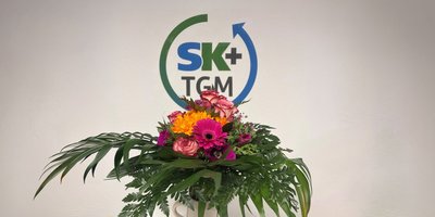 Ein bunter Blumenstrauß mit Rosen, Gerbera und anderen Blumen steht vor einer hellen Wand mit einem Logo.