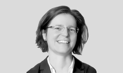 Eine lächelnde Frau mit Brille und schulterlangen Haaren trägt einen schwarzen Blazer und ein weißes Hemd.