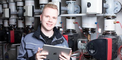 Ein junger Anlagenmechaniker vor einer Heizungsanlage, der ein Tablet hält 